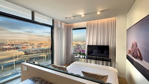 A modern hotel room with a large window offering a city view, a bed with a view, a flat-screen TV, and a small table.