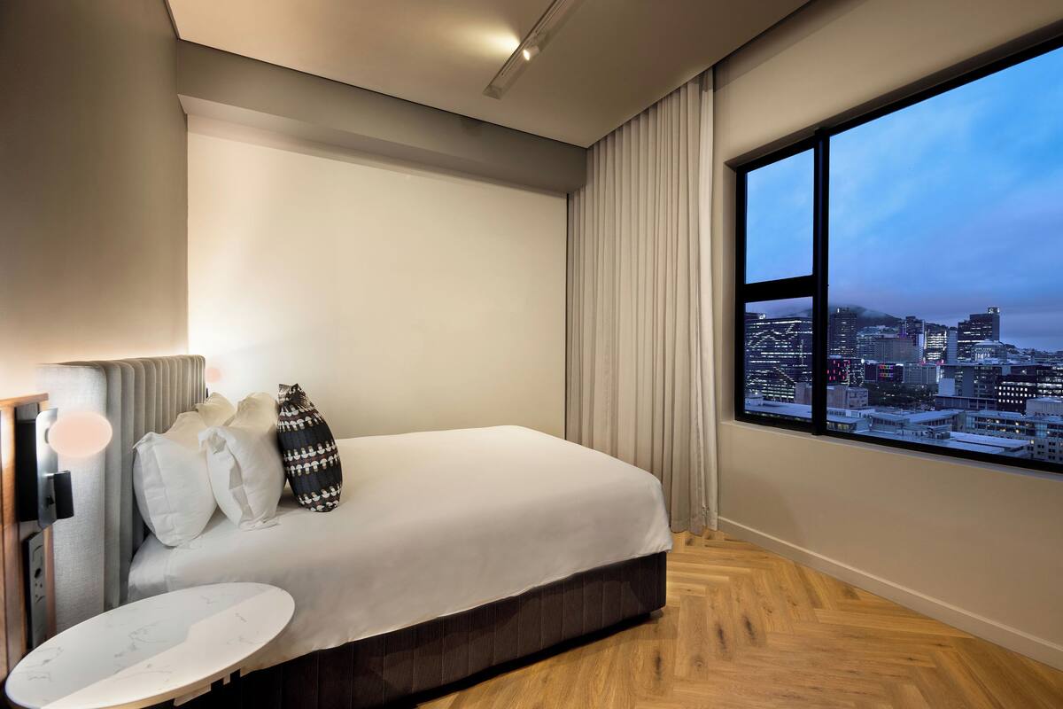 Ein modernes Hotelzimmer mit einem großen Bett, einem Nachttisch und Blick auf eine Stadtlandschaft durch ein großes Fenster.