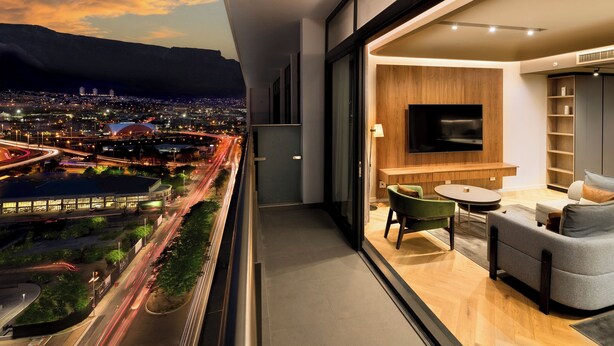A modern living room with a TV, seating, and a view of a cityscape at dusk.