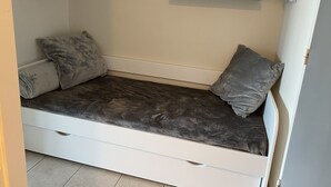 A small, single-bed room with a grey mattress, two grey pillows, and a white drawer unit. There are two shelves above the bed with various items.