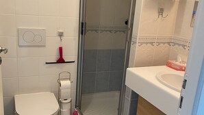 A bathroom with a toilet, a shower with a glass door, a sink, and a small shelf with toiletries.