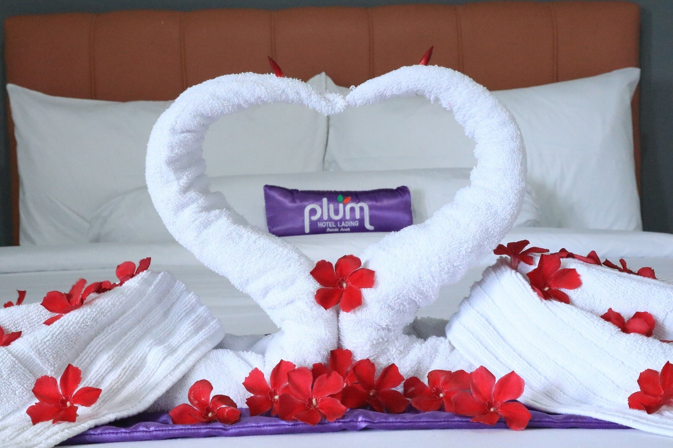 A neatly made bed with a heart-shaped towel arrangement, a purple pillow, and red flowers.
