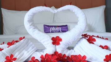 A neatly made bed with a heart-shaped towel arrangement, a purple pillow, and red flowers.