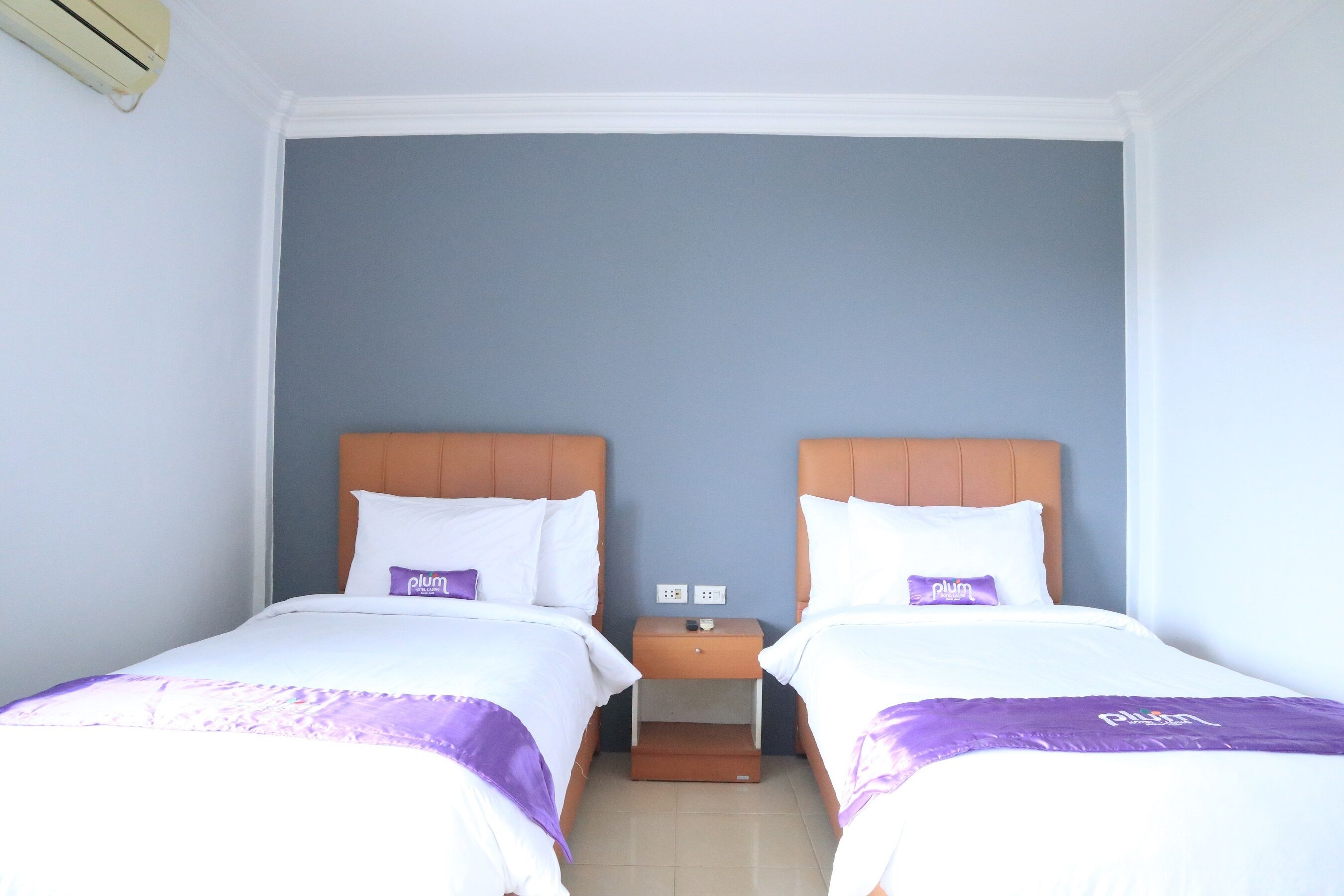 Habitación doble con dos camas, cada una con cabecero de madera, ropa de cama blanca y almohada morada. Hay una mesita de noche entre las camas, con una pequeña mesa y un enchufe.