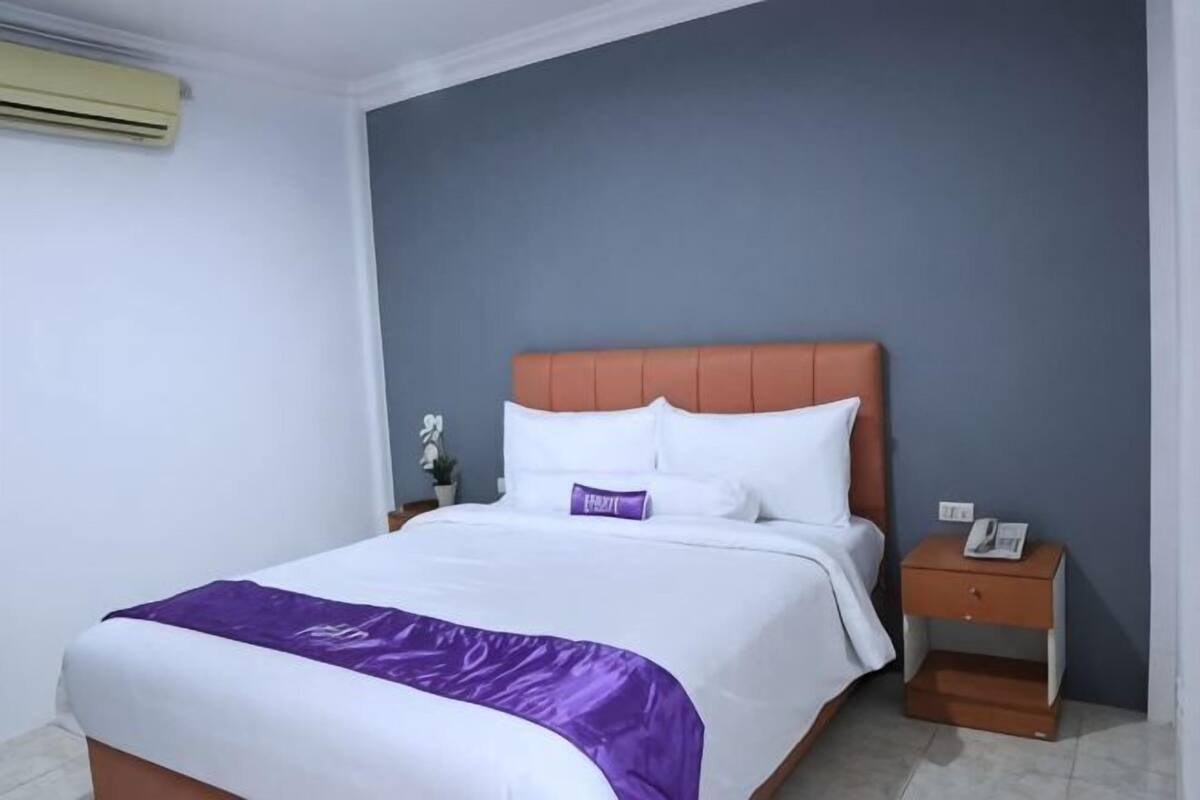 A neatly made bed with a purple throw, a wooden nightstand with a phone, and a wall-mounted air conditioner.