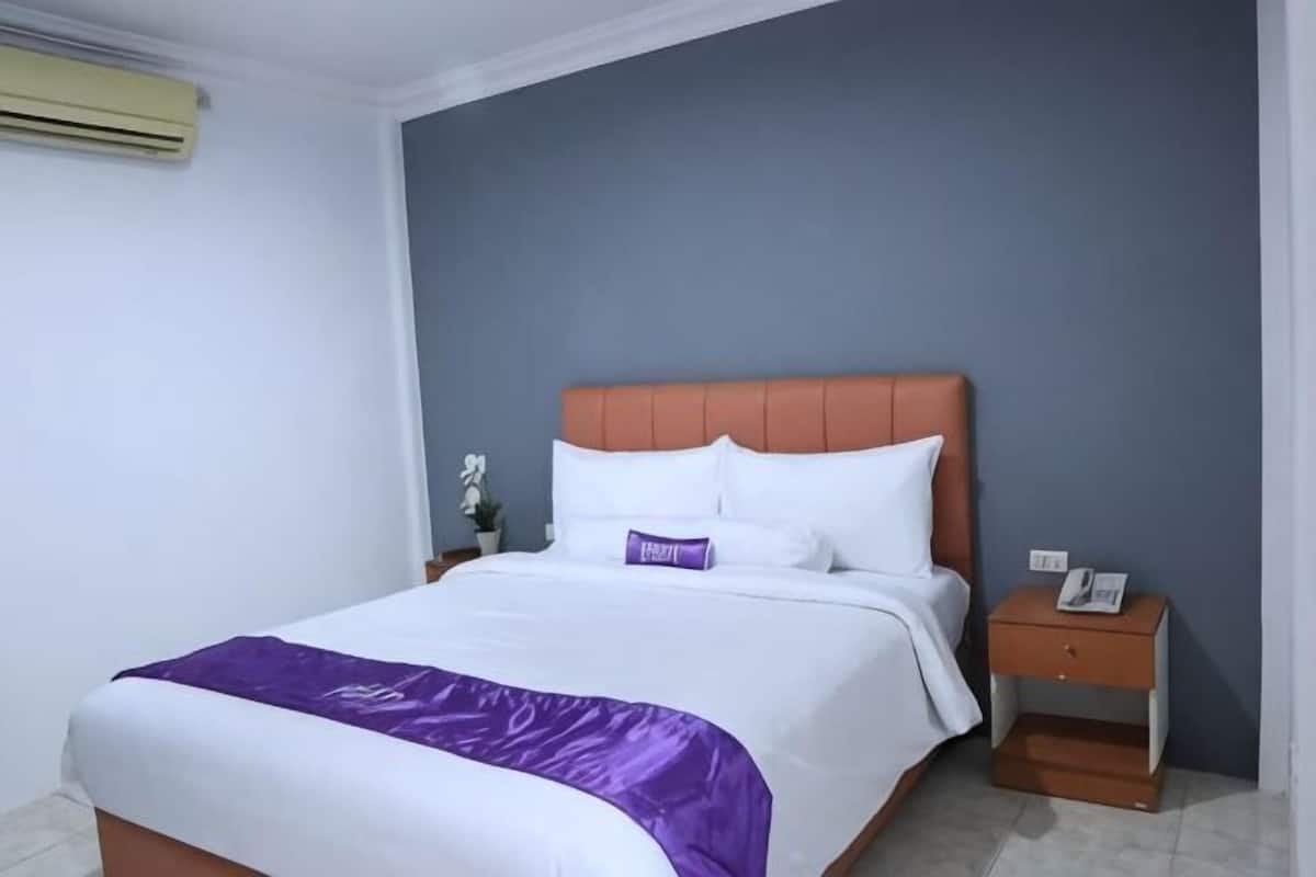 Uma cama bem arrumada com um manta roxa, uma mesa de cabeceira de madeira com um telefone e um ar condicionado fixado na parede.