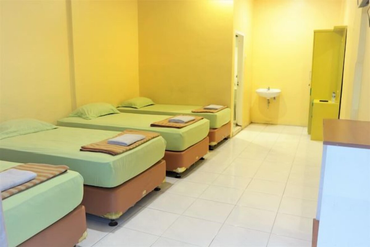 Ein Hotelzimmer mit drei Einzelbetten, jeweils mit einem Kopfkissen und einem Buch. Es gibt ein weißes Waschbecken und einen Spiegel an der Wand.