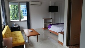 Un cuarto de hotel con un sofá amarillo, una mesa de madera, una cama con una colcha morada, un televisor montado en la pared y un ventanal con cortinas.