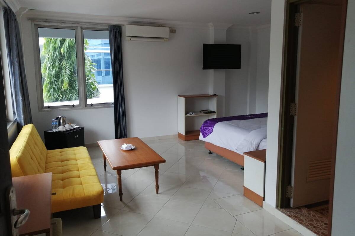 A hotel room with a yellow sofa, a wooden table, a bed with a purple blanket, a TV mounted on the wall, and a window with curtains.