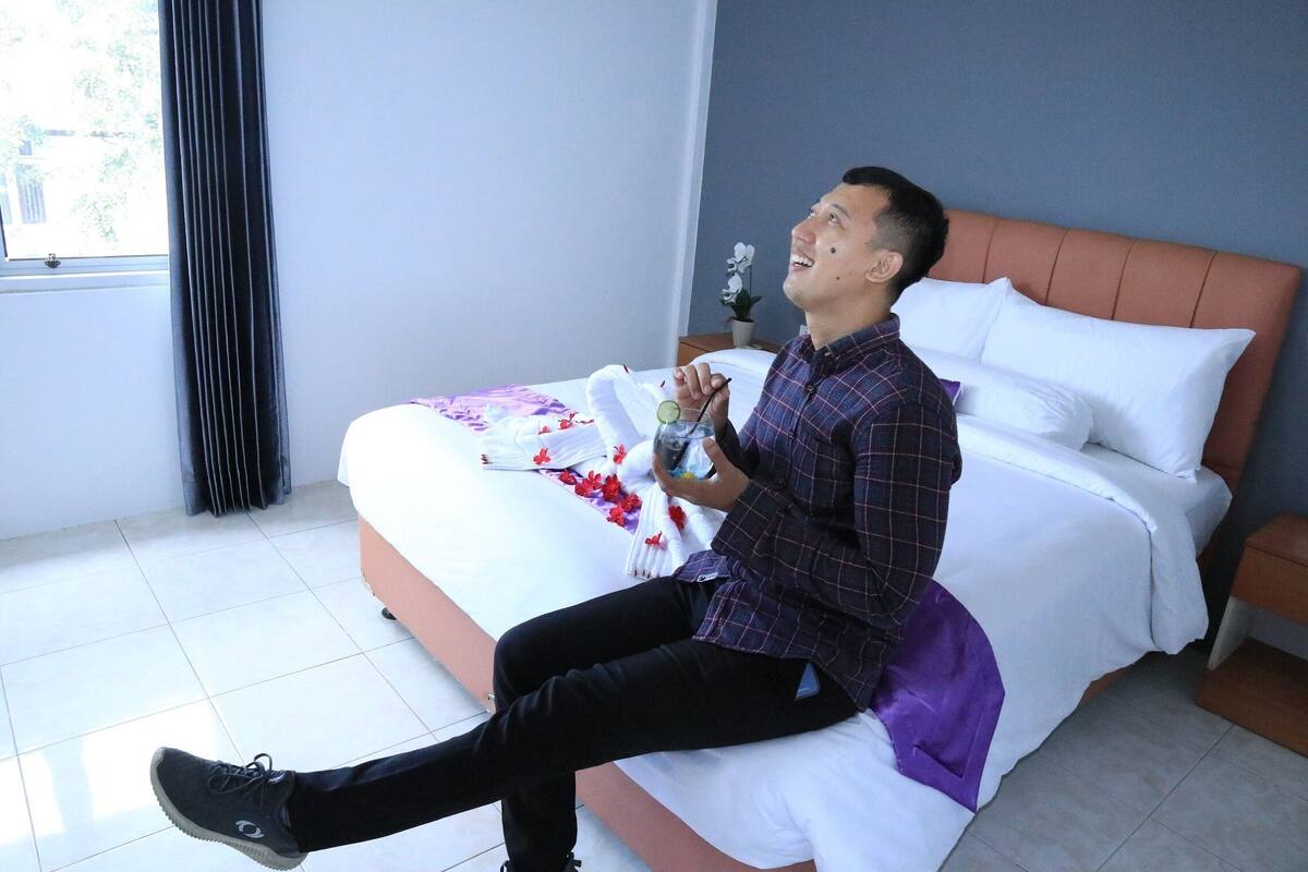A bed with white bedding and a purple blanket, a small table with a potted plant, and a window with curtains.