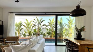 Moderni olohuone, jossa on sohva, ruokapöytä ja tuoleja. Suuresta ikkunasta avautuu näkymä trooppisiin kasveihin.