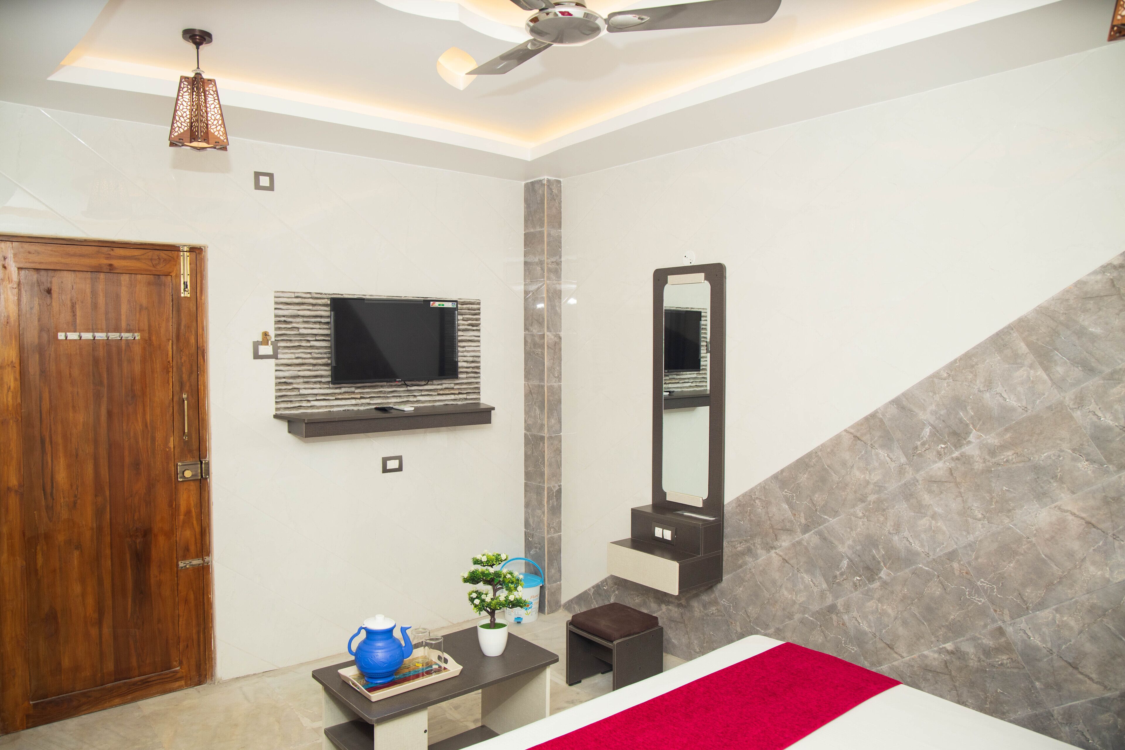 A modern bedroom with a wooden door, a flat-screen TV mounted on the wall, a large mirror, and a red accent wall.