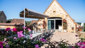 A stone building with a triangular roof, a covered outdoor dining area with chairs, and potted plants.