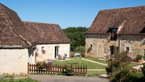 A stone building with a tiled roof, a wooden fence, and a small garden with flowers and chairs.