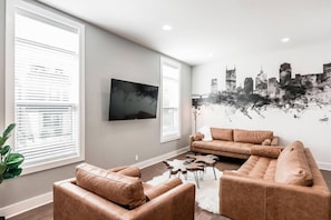 A living room with a large flat-screen TV, two sofas, a coffee table, and a cityscape mural on the wall.