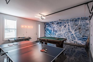 A room with a ping pong table, a pool table, and a neon sign.