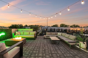 An outdoor seating area with a fire feature, string lights, and a view of the city at dusk.