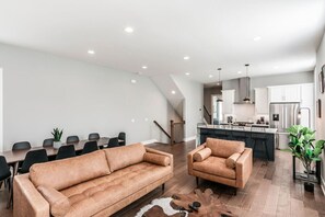 A modern living room with a sofa, armchair, dining table, and chairs. A staircase leads to the kitchen area.