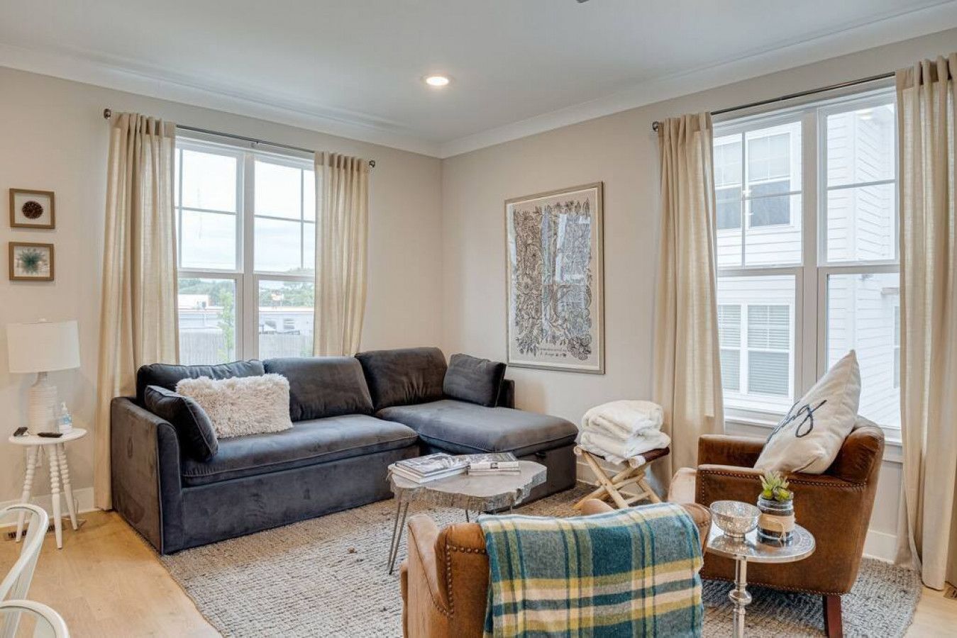 A living room with a dark grey sofa, a small round table, and a brown armchair. There is a window with curtains, a framed artwork on the wall, and a lamp on a side table.