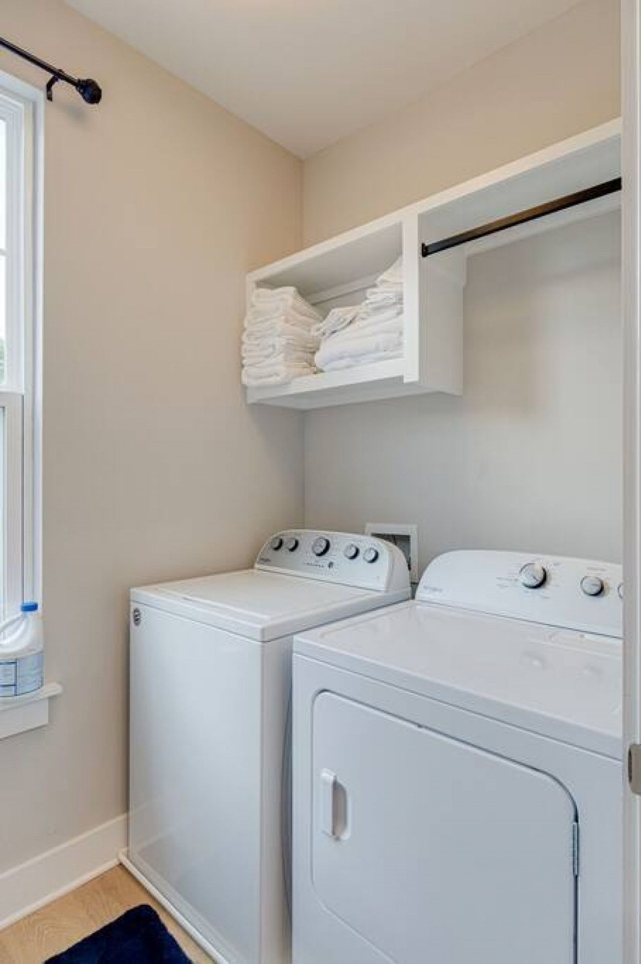 Une buanderie équipée de deux lave-linge et deux sèche-linge à chargement frontal, tous deux de couleur blanche, avec une étagère au-dessus des sèche-linge et une fenêtre sur le côté gauche.