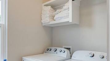 Une buanderie équipée de deux lave-linge et deux sèche-linge à chargement frontal, tous deux de couleur blanche, avec une étagère au-dessus des sèche-linge et une fenêtre sur le côté gauche.