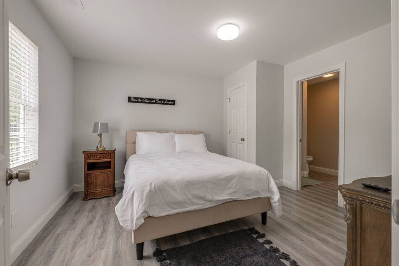 A bedroom with a bed, a nightstand, a lamp, a sign on the wall, and a door leading to another room.