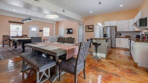 Un amplio salón con mesa de billar, una mesa de comedor con sillas, una cocina con armarios blancos y un televisor montado en la pared.