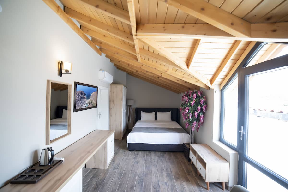 A bedroom with a wooden ceiling, a bed, a bedside table, a mirror, a window, and a wall-mounted light fixture.