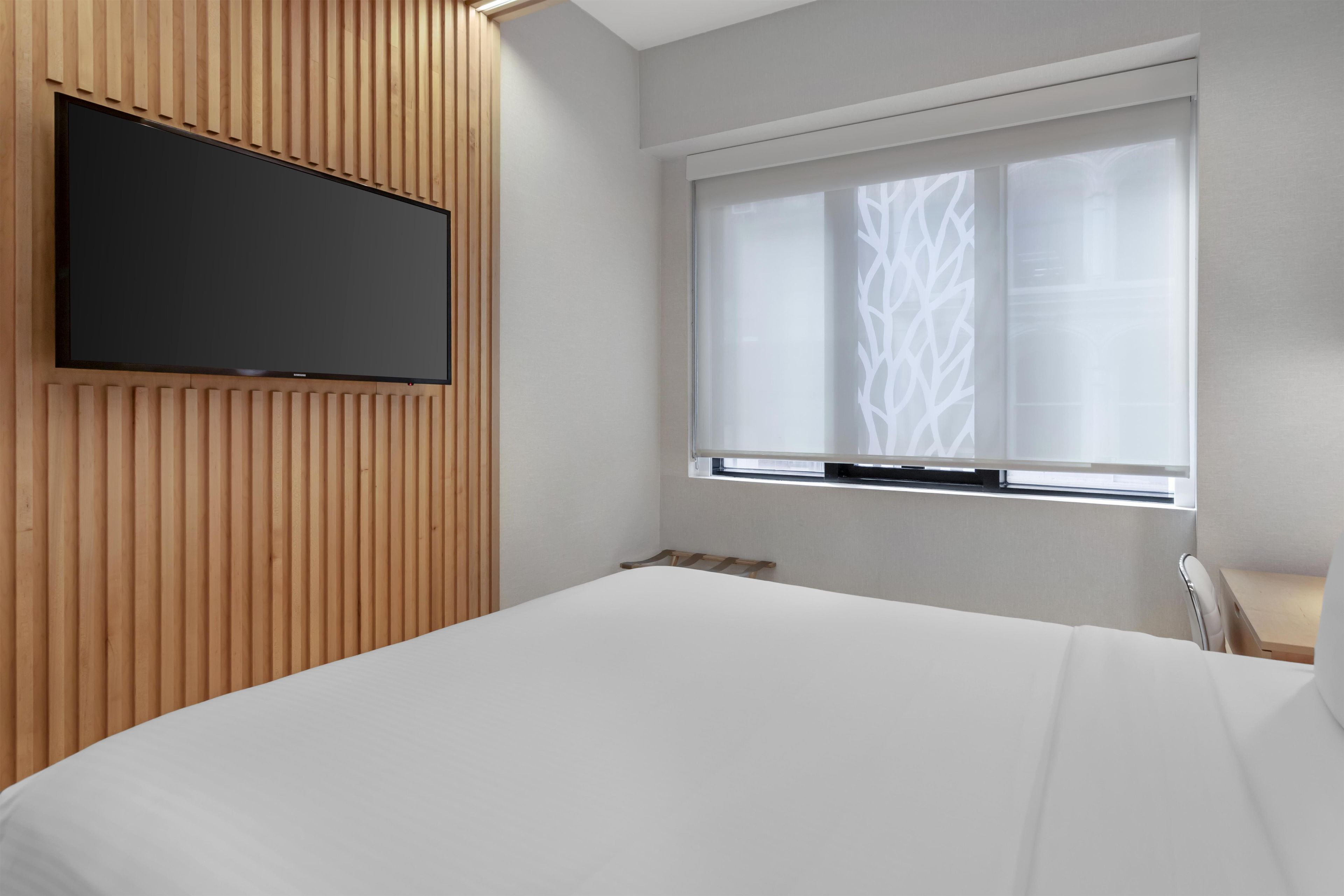 Ein modernes Schlafzimmer mit einem großen Bett, einem Wandfernseher und einem Fenster mit geometrischem Muster.