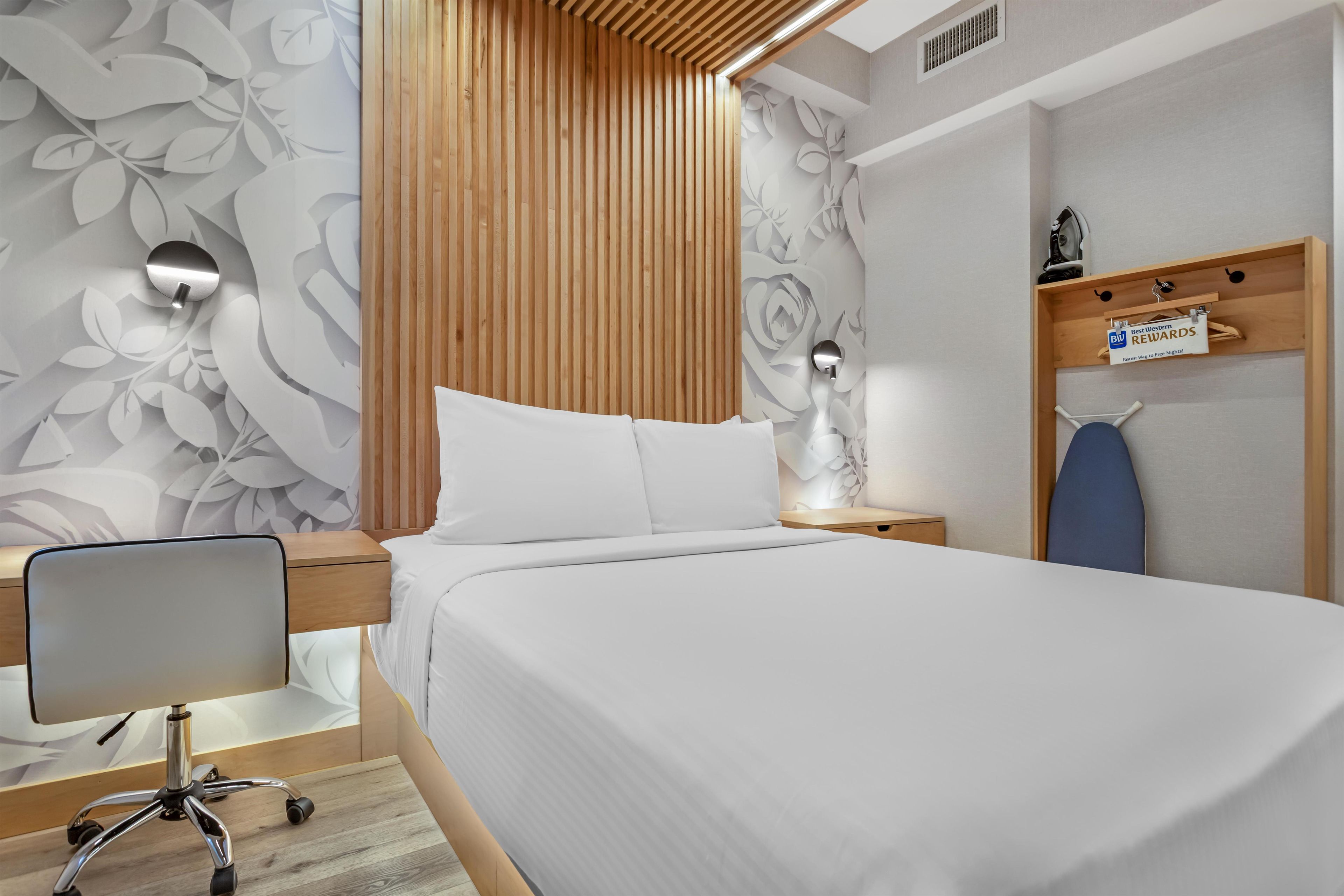 Une chambre d’hôtel moderne équipée d’un lit, d’un bureau avec une chaise, d’une lampe fixée au mur et d’une étagère en bois avec une pancarte.