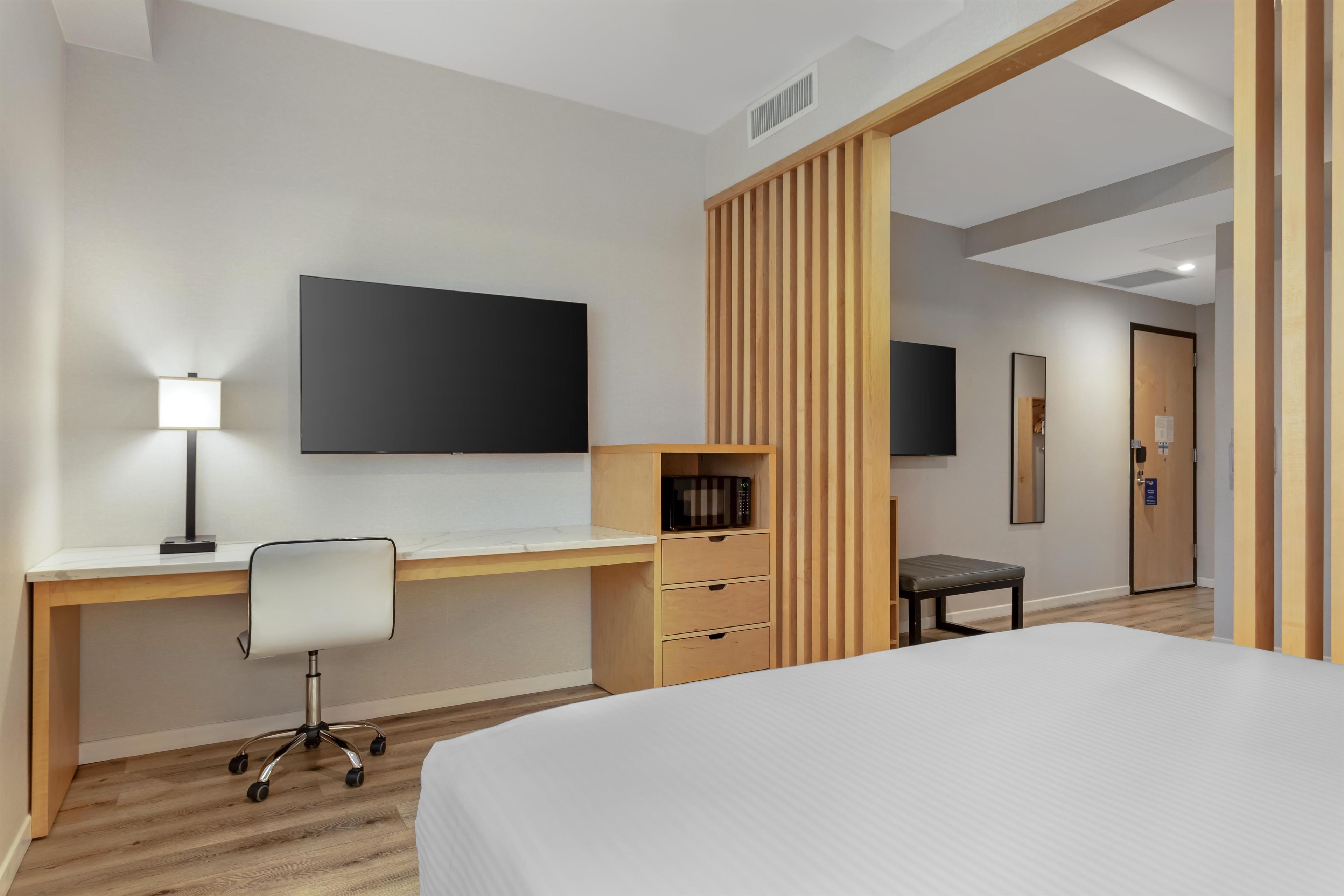 Quarto de hotel com cama, escrivaninha e cadeira, televisão de tela plana, pequena mesa com cadeira e divisória de madeira.