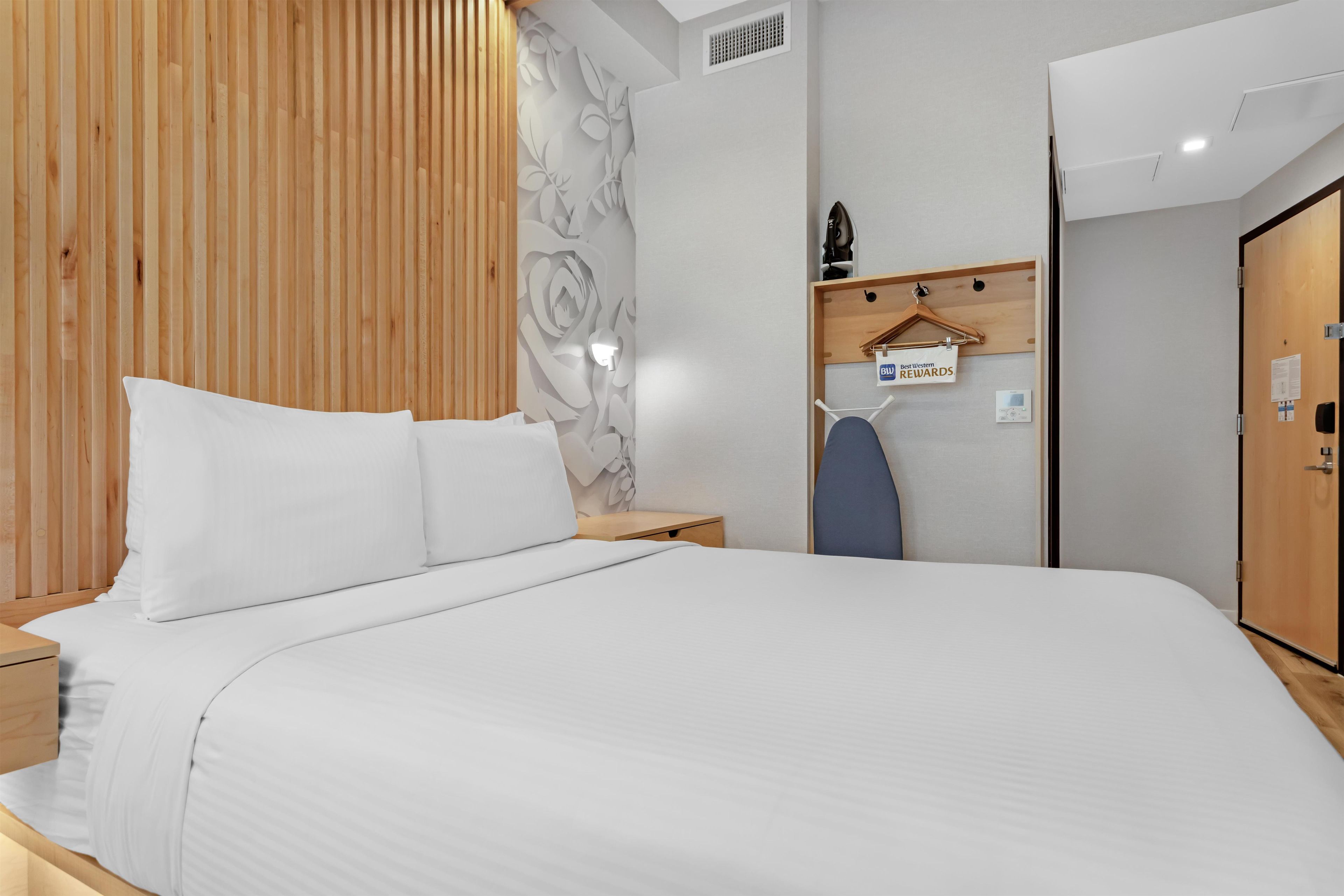 A modern hotel room with a large bed, a wooden headboard, a desk with a chair, a wardrobe, and a wall-mounted heater.