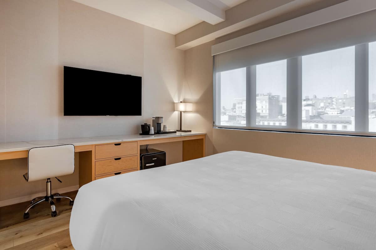 A modern hotel room with a large bed, a desk with a chair, a flat-screen TV, a lamp, and a city view through large windows.