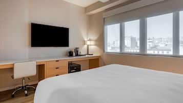 A modern hotel room with a large bed, a desk with a chair, a flat-screen TV, a lamp, and a city view through large windows.