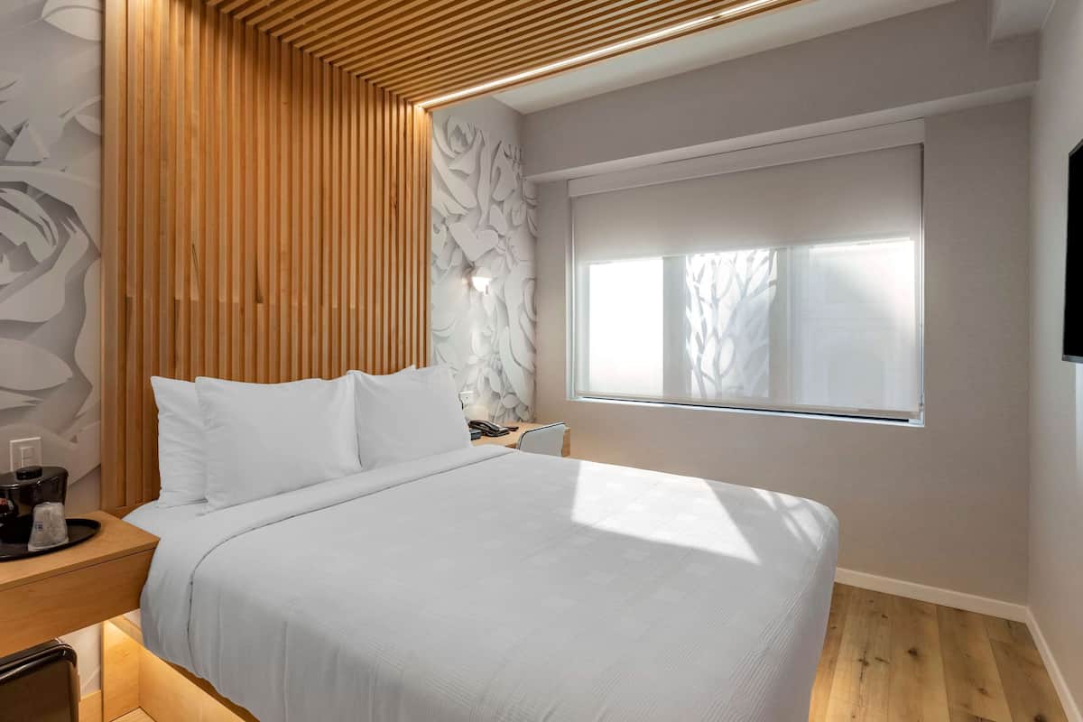 A modern bedroom with a large bed, wooden paneling, a wall-mounted light, and a window with blinds.