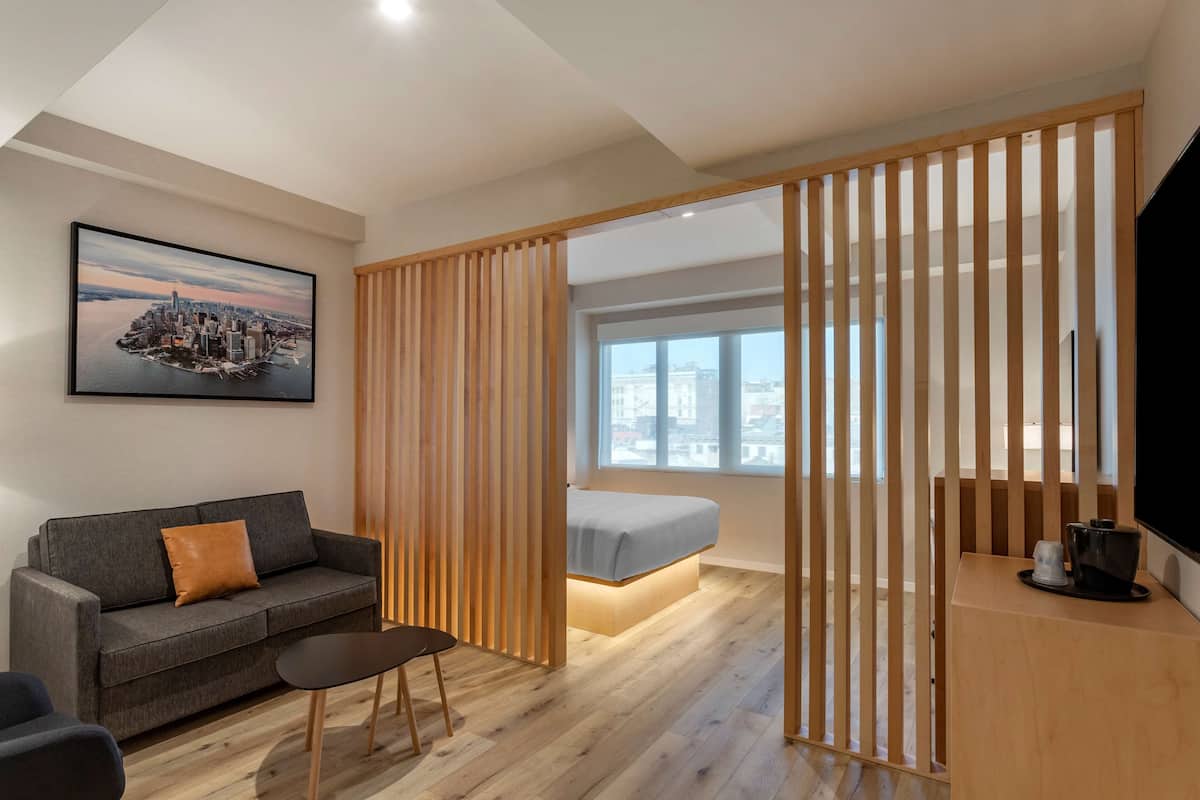 A modern hotel room with a wooden partition, a gray sofa, a small table, a bed with a light-colored headboard, a large window with a city view, and a framed cityscape painting on the wall.