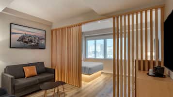 A modern hotel room with a wooden partition, a gray sofa, a small table, a bed with a light-colored headboard, a large window with a city view, and a framed cityscape painting on the wall.