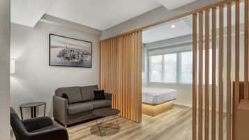 A modern hotel room with a grey sofa, a small round table, a bed with a wooden headboard, and a large window.