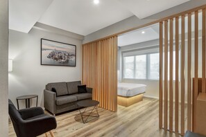 A modern hotel room with a grey sofa, a small round table, a bed with a wooden headboard, and a large window. - Best Western Plus Soho Hotel (New York)