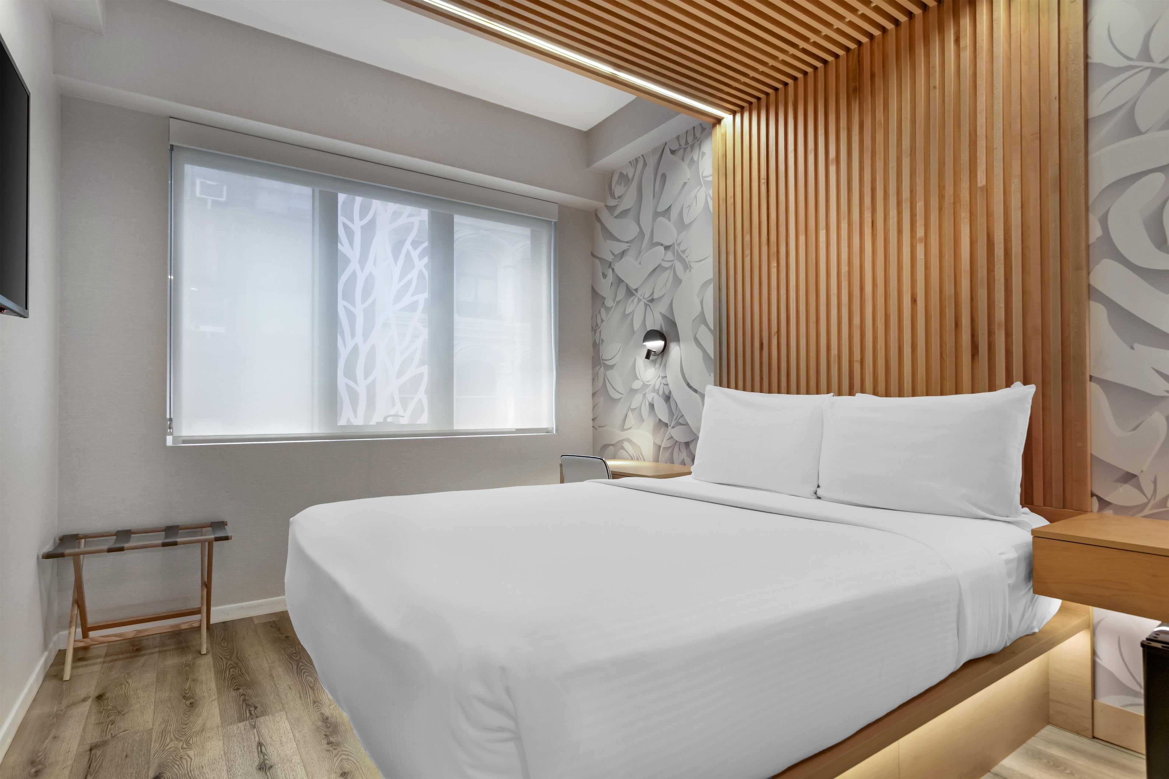 Quarto moderno com uma cama grande, uma mesinha de cabeceira, uma luminária e uma janela com cortina estampada em folhas.