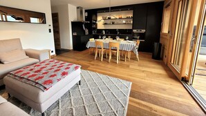 Ein modernes Wohnzimmer mit einer Couch, einem Esstisch für vier Personen und einer Küchenzeile mit schwarzen Schränken und einem Backofen.