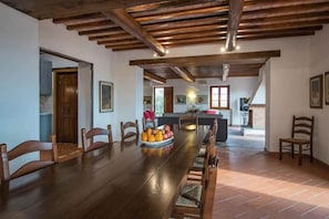Een ruime eethoek met een lange houten tafel, omringd door houten stoelen. Er staat een fruitschaal op tafel. De kamer heeft zichtbare houten balken in het plafond en een betegelde vloer.