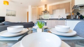 Een moderne keuken met lichte kastjes, een centraal eiland met een plant en een eetgedeelte met een glazen tafel en witte borden.