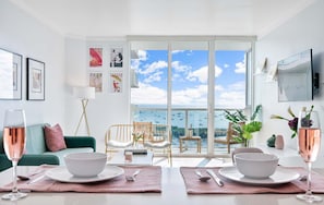 A modern living room with a dining area, large windows with a view of the sea, and various framed pictures on the walls.
