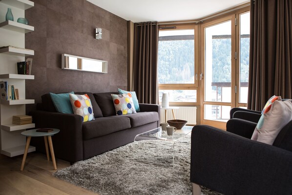 Ein modernes Wohnzimmer mit einer dunklen Couch, einem klaren Couchtisch und Blick auf eine Berglandschaft durch große Fenster.
