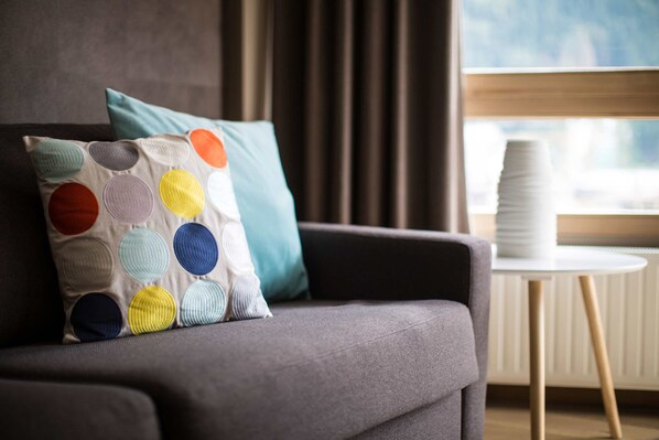 Un salon moderne avec un canapé gris, un coussin coloré, un coussin turquoise et une pile d’assiettes blanches sur une petite table ronde.