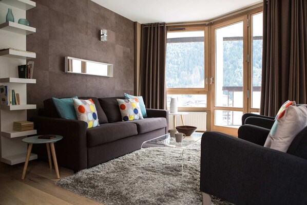 A modern living room with a dark sofa, colorful pillows, a glass coffee table, and a view of a snowy landscape through large windows.