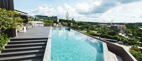 A rooftop infinity pool with clear blue water, surrounded by a wooden deck and greenery.