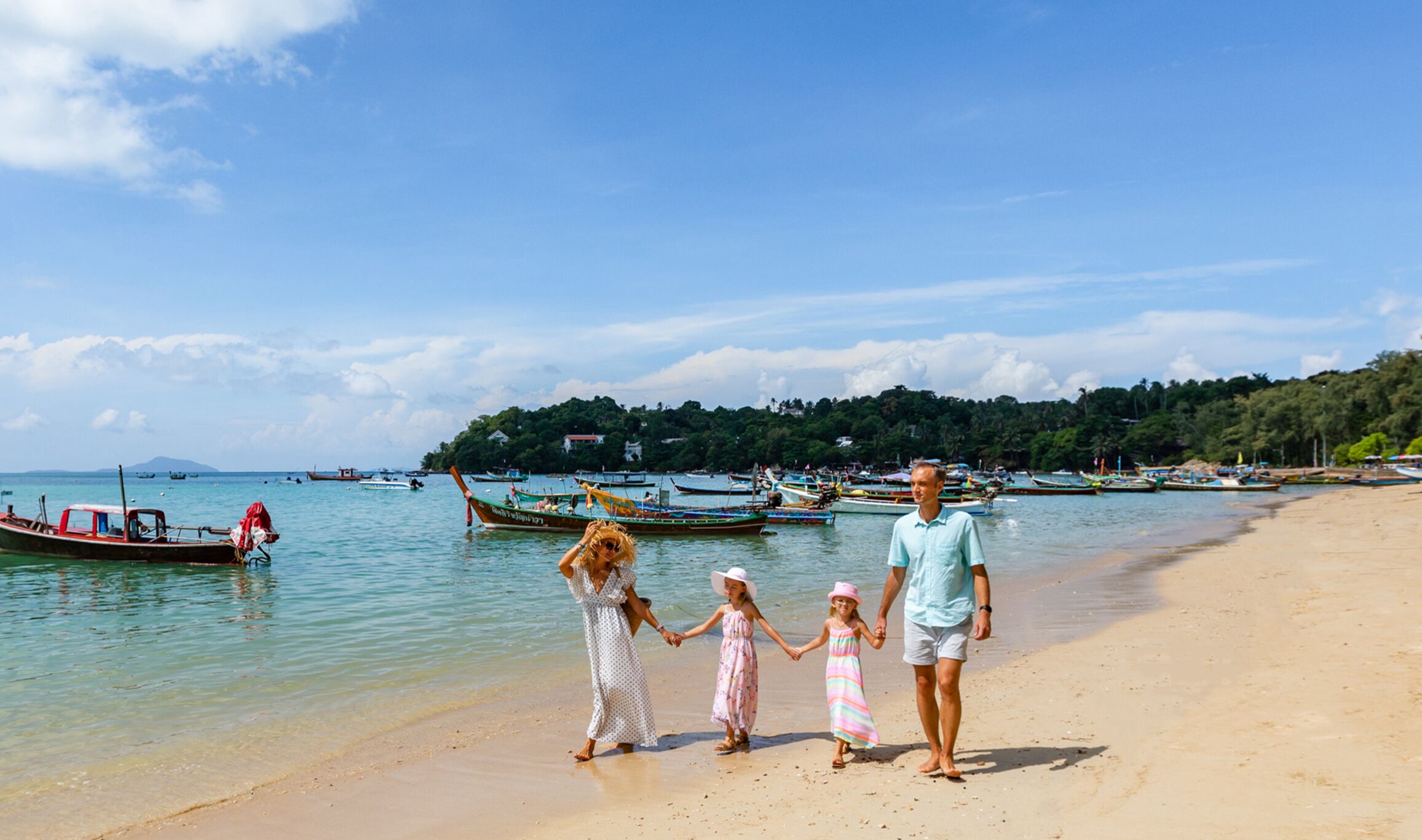 Una playa con una familia caminando por la orilla, barcos en el agua y una colina boscosa al fondo.