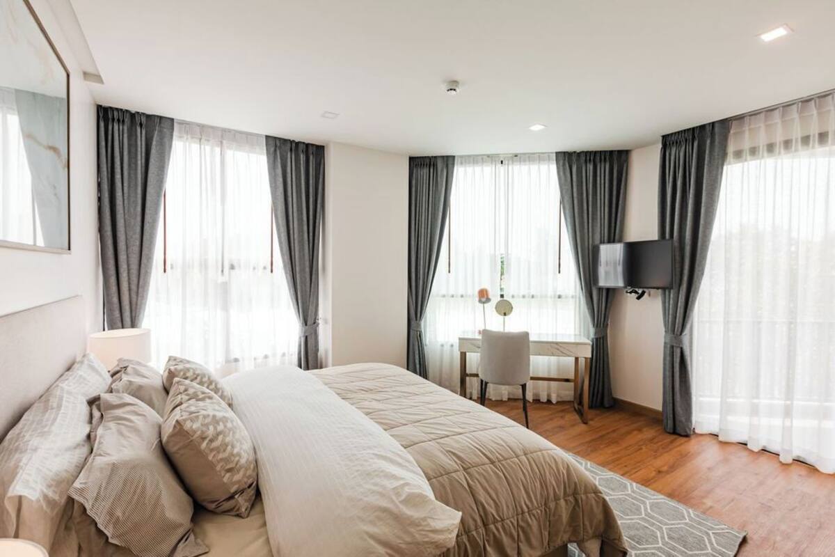 A modern hotel room with a large bed, a desk with a chair, a television, and large windows with curtains.
